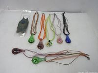 Eight colorful handmade hand blown glass pendants with cloth necklaces arranged on a white surface, showing color variations and shapes.