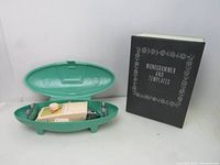 Singer buttonholer tool in open green case alongside a black Monogrammer and Templates book standing upright.