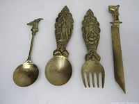 Four brass utensils on white background including letter opener, fork, two spoons with decorative Asian styled handles
