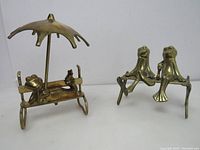 Both brass frog figurines together, showing single frog under umbrella on left and frog couple on right on benches.