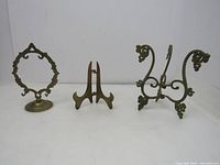 Three vintage brass items displayed on white surface: round ornamental picture holder, foldable three-legged easel, ornate decorative brass stand.