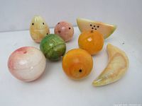 Overview of eight carved marble/alabaster fruits including watermelon slice, banana, pear, orange, apple and others, shown on white surface
