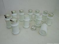 Front view showing 11 vintage milk white small spice jars with silver lids, one jar missing lid, varying presence of plastic inserts with sprinkling holes.