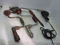 Tools arranged with Wen soldering iron, vintage electric stick soldering iron with wood handle, and two vintage hand drills with wood ends and cranks on a white surface.