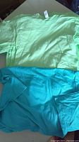 Photo of two American Apparel ladies T-shirts, one mostly light green and another turquoise blue, laid flat showing front sides.