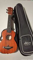 Aklot AKS21 wooden ukulele with nylon strings next to the black padded carrying case with Aklot logo.