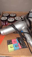 Silver Revlon hair dryer with power cord, visible buttons, and colored hair clips in front.
