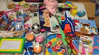Overview of many toys including a pink plush pig, dartboard game, Nerf blaster, packaged small toys, books, squishy ball toys, and more.