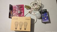Various light bulbs including a 3-pack of UV black light LED bulbs, a Philips LED bulb pack, pink small bulbs, loose bulb wired to a socket, and socket cord with plug.