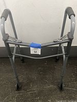 Front angle view of a used grey metal hand walker with grey padded handles, two small front wheels, and rubber-tipped rear adjustable legs on a floor surface against a wall.