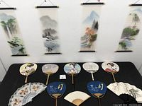 Four Japanese hanging scrolls depicting nature scenes including trees, mountains, water, and foliage hung on a wall above a black table with nine handheld fans.