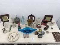 Photo shows assorted vintage and decorative items arrayed on a white surface. Items include glass bottles, ceramic pitchers, figurines, plaques, and small collectibles.