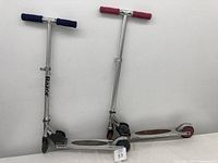 Pair of two silver Razor kids scooters, one with blue grips and one with red grips, leaning against a white wall.