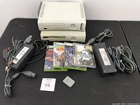 Two white Xbox 360 consoles shown with power supplies, cords, and three Xbox 360 game cases plus a memory card on table.