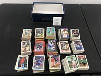 Wide view of multiple stacks of baseball cards arranged in front of an empty shoe box on a black surface, showing the diversity and quantity of cards in the lot.