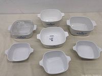 Seven white CorningWare baking dishes with blue cornflower pattern, one with clear glass lid, shown from top and angle views.