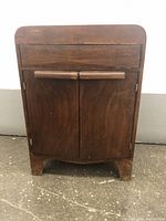 Front view of vintage wooden sewing machine cabinet with two doors and drawer above. Shows surface wear and wooden handles on doors.