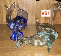 Front view of two art glass fish sculptures on wooden surface, one blue upright and one clear aqua-tinted lying down.