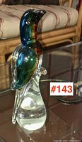 Front left side view of an 8.5 inch tall art glass bird with green and blue tones sitting on a solid clear glass base, positioned on a glass table.
