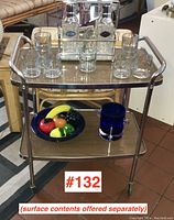 Full view of the MCM chrome bar cart displaying multiple glass decanters, glasses on top shelf, and a bowl with decorative fruit on bottom shelf.