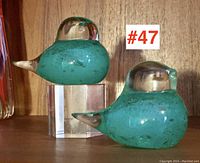 Pair of turquoise blue art glass bird figurines displayed on a wood surface, showing overall shape and size with one bird elevated on a clear stand.
