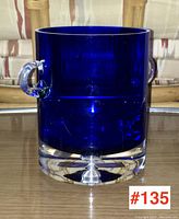 Front view of cobalt blue glass ice bucket with clear glass handles and thick clear base on a reflective surface.