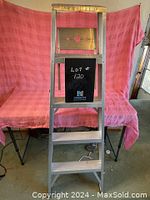 Front view of four step aluminium ladder with some tools placed beside it and pink cloth backdrop.