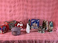 Wide shot showing various Christmas-themed decorative houses, holiday containers, and boxed collectibles arranged on a pink cloth backdrop.