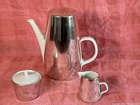 Photo showing the coffee pot with removable shiny silver-colored warmer top, creamer, and sugar bowl against a pink fabric background.
