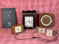 Five vintage clocks on pink backdrop: large wooden mantle clock, square wooden wall clock, rectangular wood alarm clock, and two small pink electric alarm clocks.