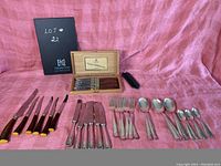 Full view of assorted cutlery laid out on cloth background including boxed Laguiole knife set, various knives, forks, big spoons, and teaspoons.