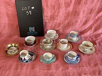 Photo showing all 10 assorted demitasse coffee cups and matching saucers in different styles, colors, and sizes, arranged on a reddish-pink fabric background.