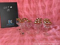 12 gold rimmed goblets arranged in two groups of 6, showing the liqueur glasses and wine goblets on a pink cloth background with a black placard labeled 'LOT 19'.