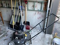 Photo of the Yard Machines lawn mower and assorted shovels in a garage setting on concrete floor, showing all items together.