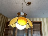 Front angle view of the chandelier hanging from the ceiling, glowing light bulb in center, showcasing the scalloped yellow stained glass petals with textured metal trim.
