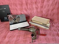 Front view of the manual meat slicer, kitchen scale, and meat grinder arranged on a pink cloth background.