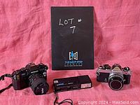 Photo showing the lot of two Nikon cameras and one Keystone Everflash 300 camera on a pink cloth background, with a black box labeled 'Lot #7'