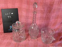 Photo showing the set of three hand cut crystal pieces against a pink backdrop: wine decanter with stopper, ice bucket with handles, and lidded jar.