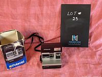 Vintage Polaroid Sun 600 LMS instant camera displayed next to its original worn box and a black cardboard box with 'LOT 23' written on it.