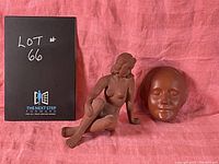 Photo showing two reddish-brown sculptures on pink fabric: a seated female nude figurine and a separate mask resembling L'Inconnue de la Seine.