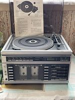 Front view of the Sears turntable unit with cassette player and AM/FM radio. Dust cover is open, instruction manual displayed behind the turntable.