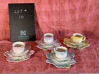 Four flower-shaped tea cups each with matching flower petal saucers in pink, purple, blue, and yellow, arranged on a red cloth background.