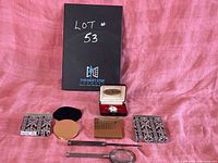 Vintage collection items arranged on pink fabric including cigarette cases, compact, letter opener, magnifying glass, and pocket watch