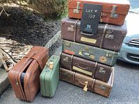 Front and side view of the stacked vintage steamer trunks with brown and green suitcases placed adjacent on the ground outside.