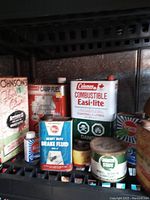 Shelf displaying a collection of vintage fuel, brake fluid, and oil tins including Coleman combustible fuel, heavy duty brake fluid, camp fuel, and others.