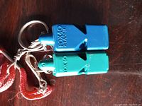 Two Fox 40 Classic whistles, blue and green, attached to red lanyards with metal keyrings and clips, displayed on wood surface.