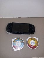 Photo shows Sony PSP handheld console with screen and control buttons. Two Universal Media Disc (UMD) game discs placed below it labeled LittleBigPlanet and Daxter.