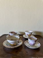 Four porcelain teacups and matching saucers arranged on a wooden surface with distinct floral designs and gold trim.