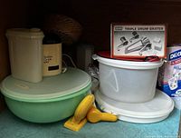 Large plastic mixing bowls, citrus juicer, triple drum grater box, Salton milk warmer, and other kitchen containers inside cabinet