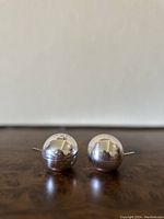 Pair of small spherical plated sterling silver salt and pepper shakers with thin side handles and perforated tops, shown from a slightly angled front view on a brown surface.